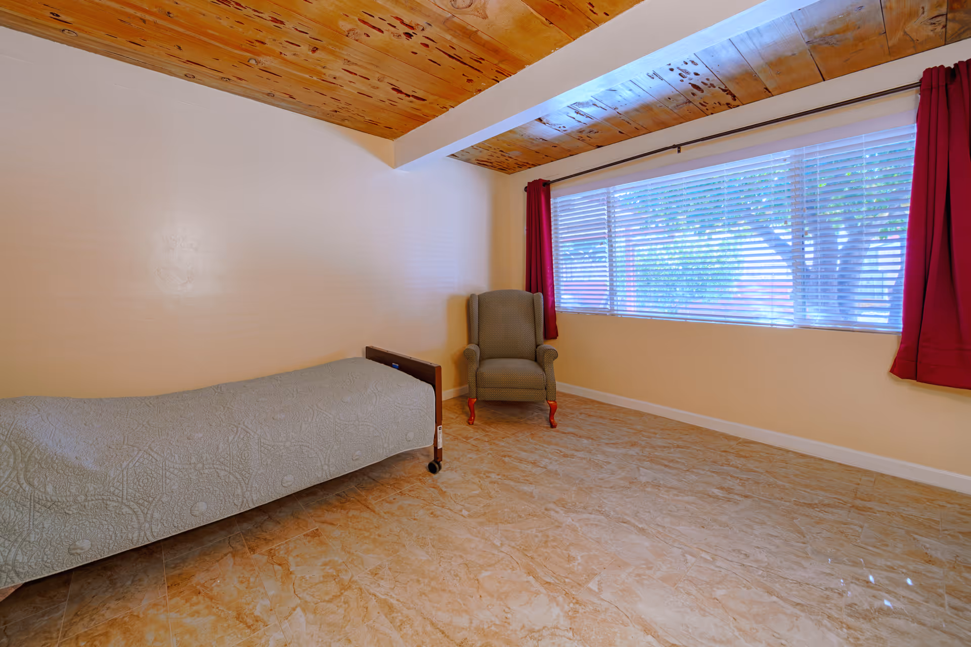 A simple bedroom with a single bed covered with a light gray patterned blanket, a green upholstered armchair with red legs, a large window with white blinds and red curtains, beige walls, a wooden ceiling, and a tiled floor.