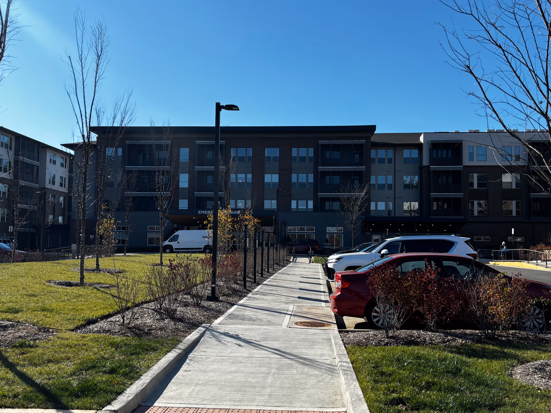 View of a modern senior living facility building with multiple floors, a sidewalk leading to the entrance, parked cars on the right, and landscaped grass and trees on both sides under a clear blue sky.