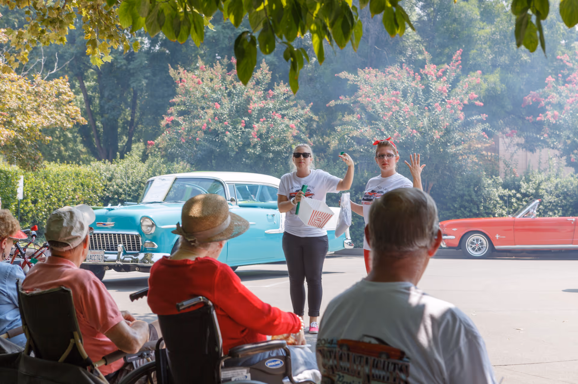 A group of elderly people sitting outdoors, some in wheelchairs, watching two women standing and engaging with them. Behind the women, there are two vintage cars, one turquoise and one red, parked on a sunny day with trees and flowering bushes in the background.