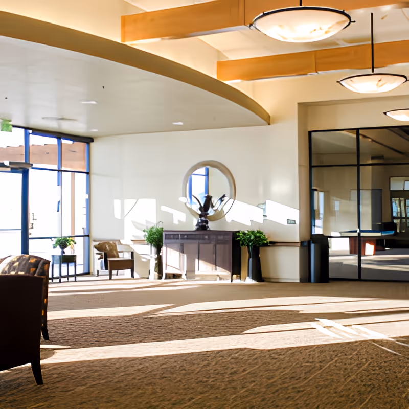 Bright and spacious interior common area with large windows letting in natural light, a round mirror above a wooden cabinet, potted plants, chairs, and modern ceiling lights.