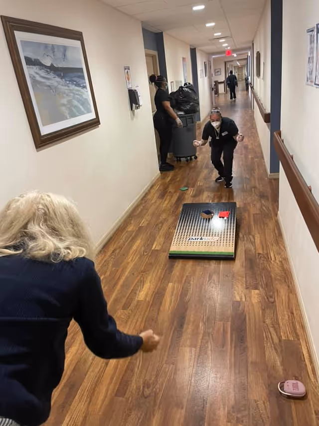 Two women playing a game of cornhole in a hallway with wooden flooring inside a senior living facility. One woman is throwing a bean bag while the other is crouched near the cornhole board. A third person is seen in the background near a trash bin. The hallway has framed artwork on the walls and handrails on both sides.