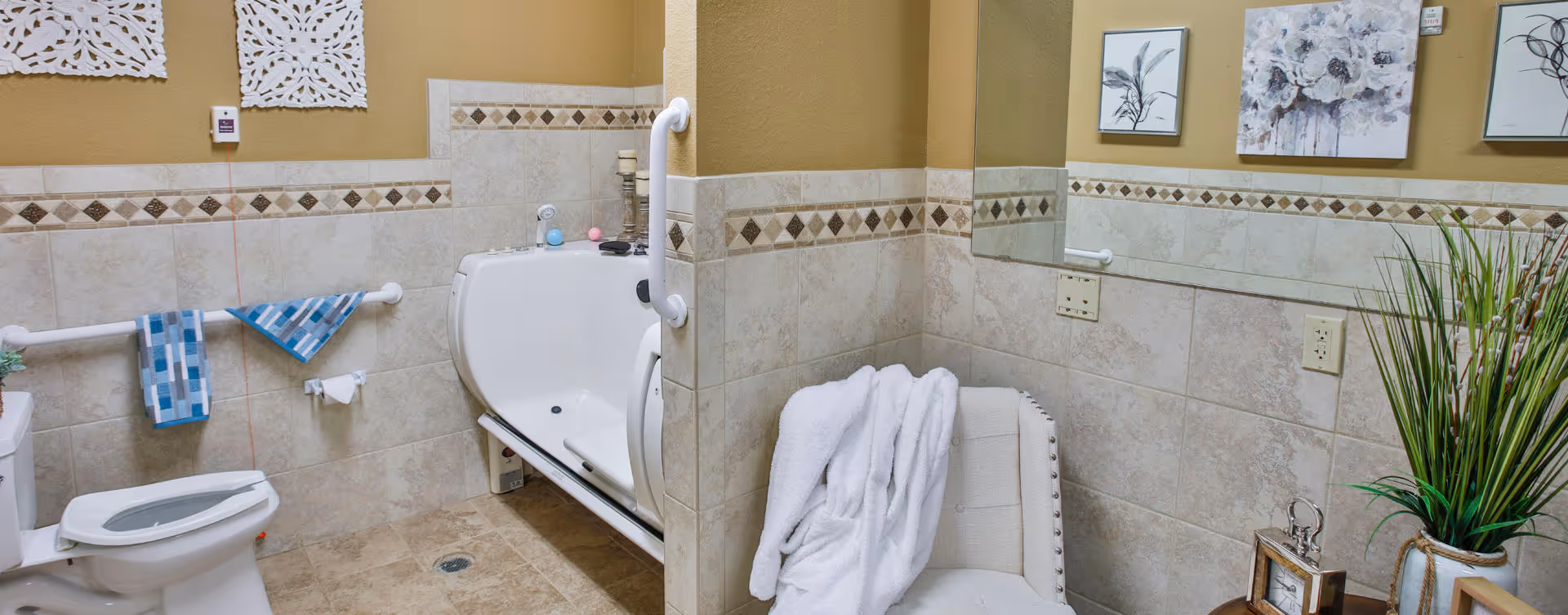A bathroom with beige tiled walls and floor, featuring a white toilet with the seat up, a walk-in bathtub with grab bars, a white chair with a white towel draped over it, decorative wall art, and a tall green plant in a pot on a small wooden table.