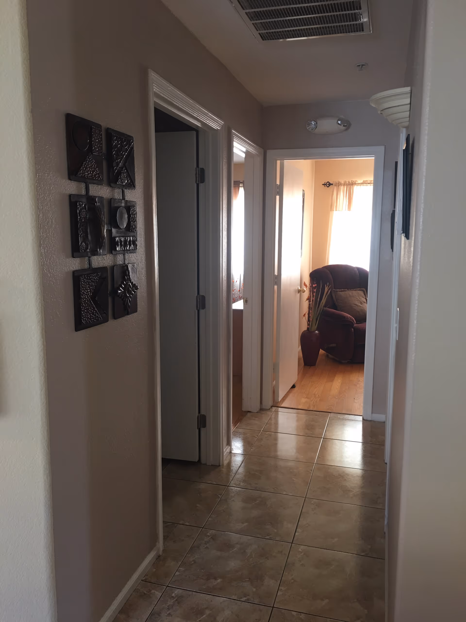A hallway with beige walls and tiled floor leading to a room with wooden flooring and a maroon armchair. There are two open doors on the left side of the hallway and decorative wall art on the left wall.