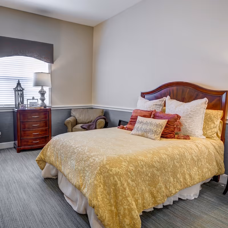 A tidy bedroom in a senior living facility featuring a wooden headboard bed with a yellow patterned comforter, several pillows, a nightstand with a lamp, and an armchair by a window.