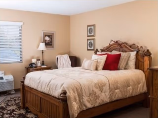 A cozy bedroom with a wooden bed frame and a beige patterned comforter. The bed is adorned with multiple pillows, including a prominent red one. There is a wooden nightstand with a lamp, a phone, and some small items next to the bed. The walls are painted a warm beige color and decorated with framed pictures. A window with white blinds is visible on the left side, and a patterned rug is partially visible on the floor.