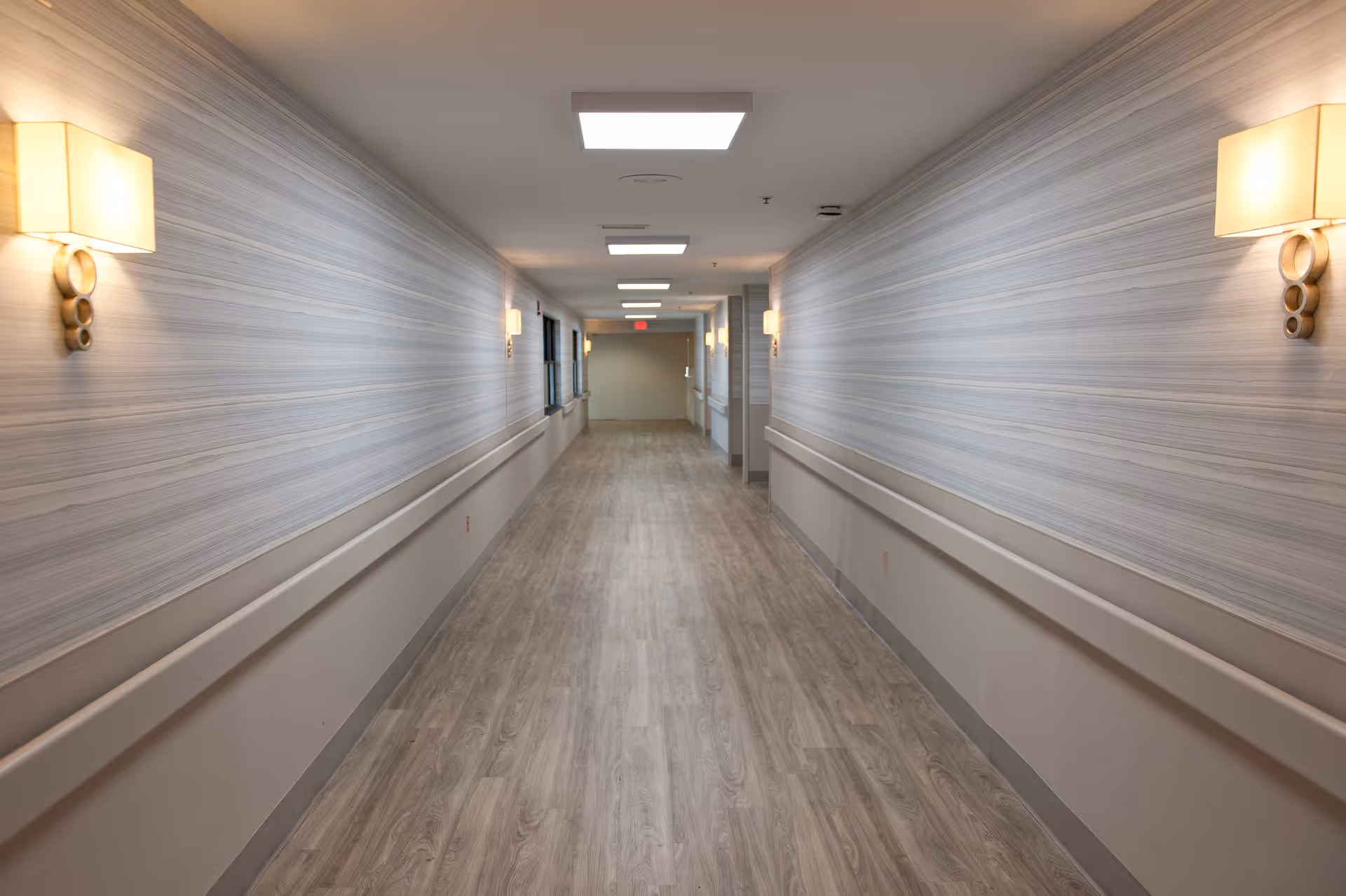 Well-lit empty hallway with wood-look flooring, handrails, and wall sconces in a senior living facility.