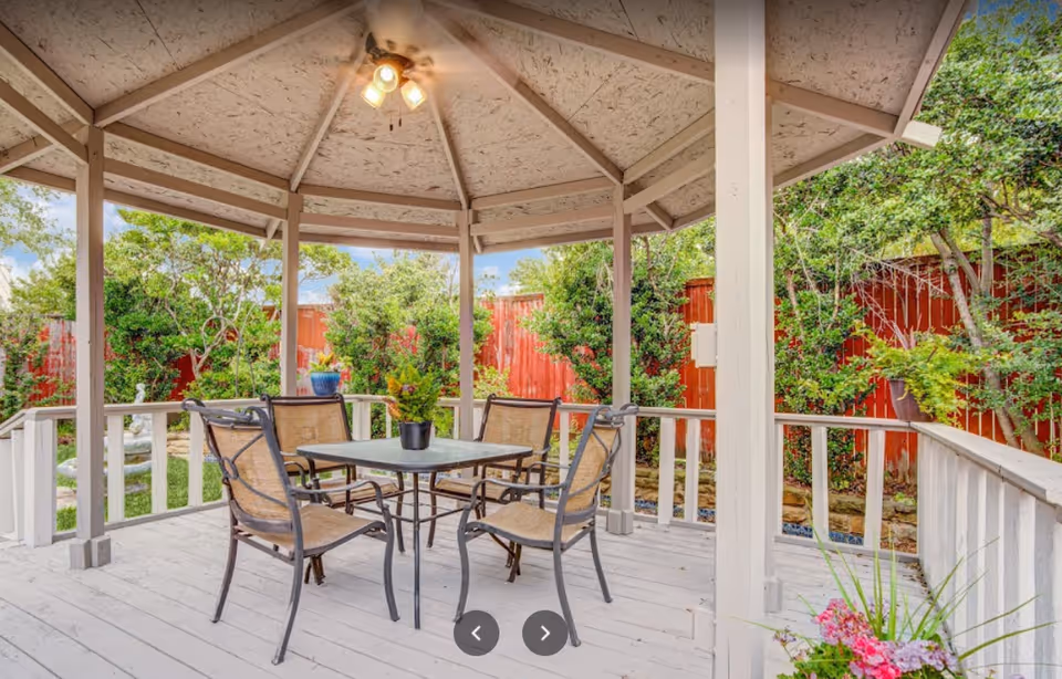 A covered outdoor gazebo with a ceiling fan and light fixture, containing a square table with four chairs. The gazebo is surrounded by greenery and a red wooden fence, with potted plants and flowers visible around the area.