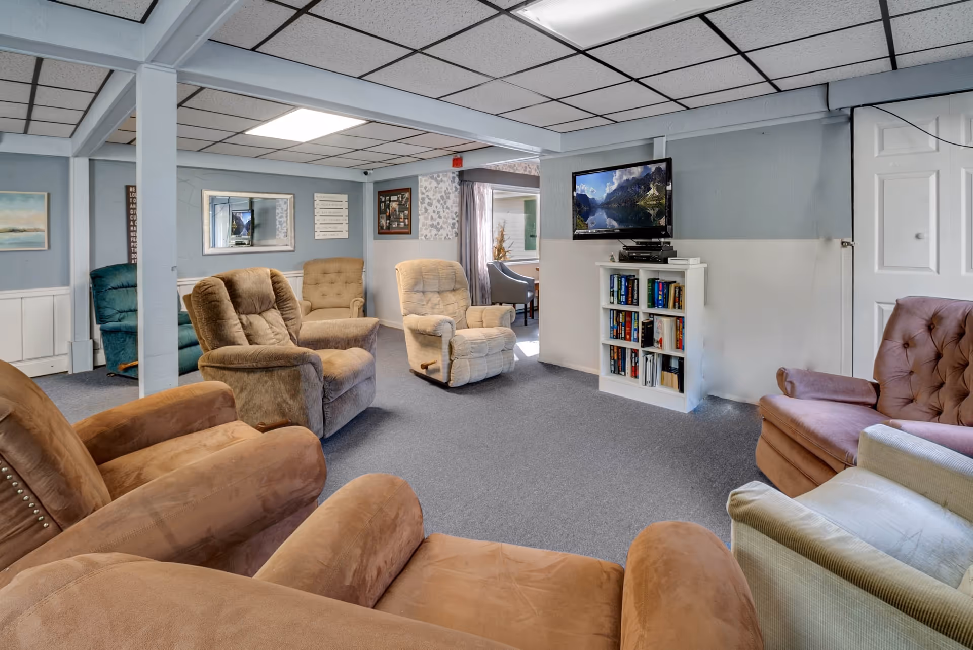 A cozy living room area with multiple cushioned recliner chairs arranged in a circle. The room has a gray carpet, light blue walls with white wainscoting, and a drop ceiling with fluorescent lighting. A flat-screen TV is mounted on the wall above a small white bookshelf filled with books. There are framed pictures and artwork on the walls, and a window with curtains is visible in the background.