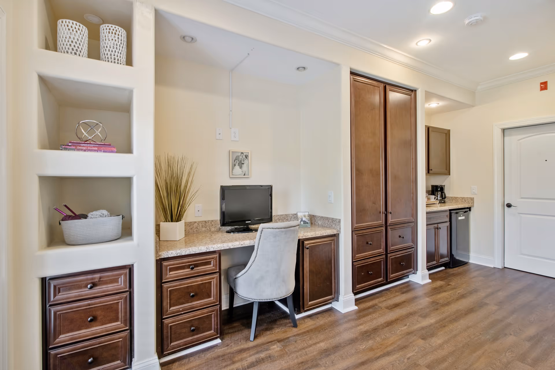 Interior view of a room with built-in wooden cabinets and drawers, a small desk area with a computer monitor and a gray upholstered chair, decorative items on shelves, and a kitchen area with a coffee maker and mini fridge in the background. The floor is wood, and the walls are light-colored with recessed lighting in the ceiling.