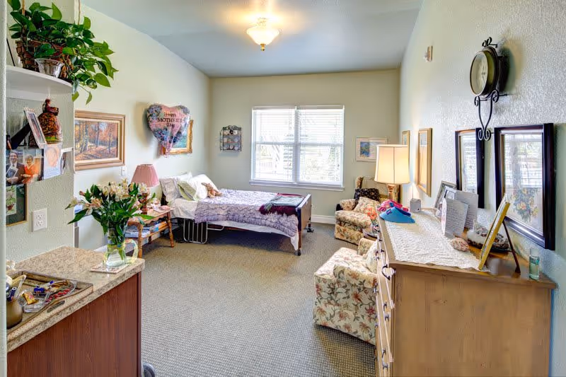 A bright and cozy assisted living bedroom with a single bed near a window, floral armchairs, a wooden dresser with framed pictures, a wall clock, and various decorations including a heart-shaped balloon and plants.