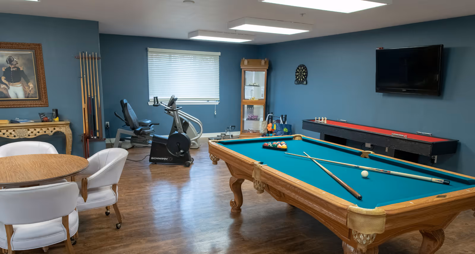 A recreational room with a pool table, shuffleboard table, exercise bike, round table with four white chairs, a dartboard on the wall, and a window with blinds. The walls are painted blue and there is a framed picture of a dog dressed in historical military attire.