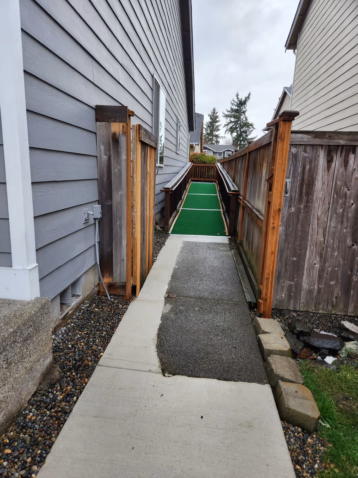 Narrow exterior walkway between two houses leading to a raised wooden ramp with green turf and handrails.