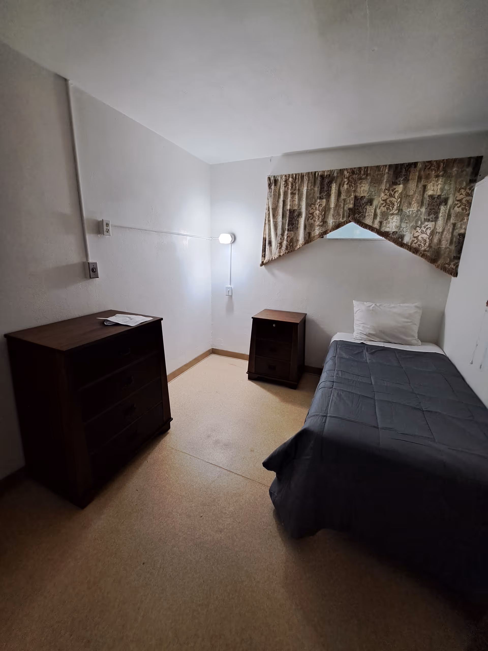 Small single-occupancy bedroom with a single bed, nightstand, dresser, wall lamp, and a curtained window.