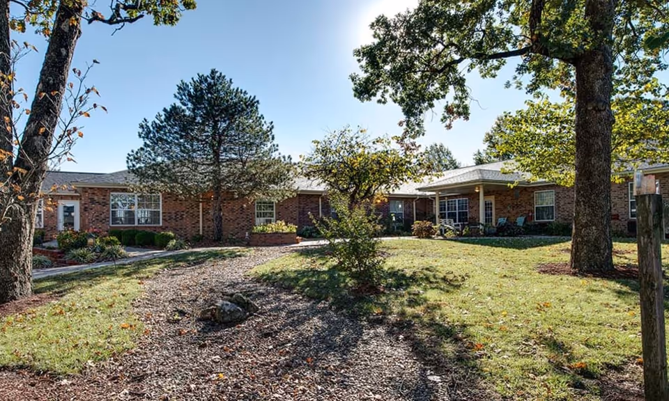 Outdoor view of a senior living facility with a brick building surrounded by trees and a grassy area with a gravel pathway leading to the entrance under a clear blue sky.