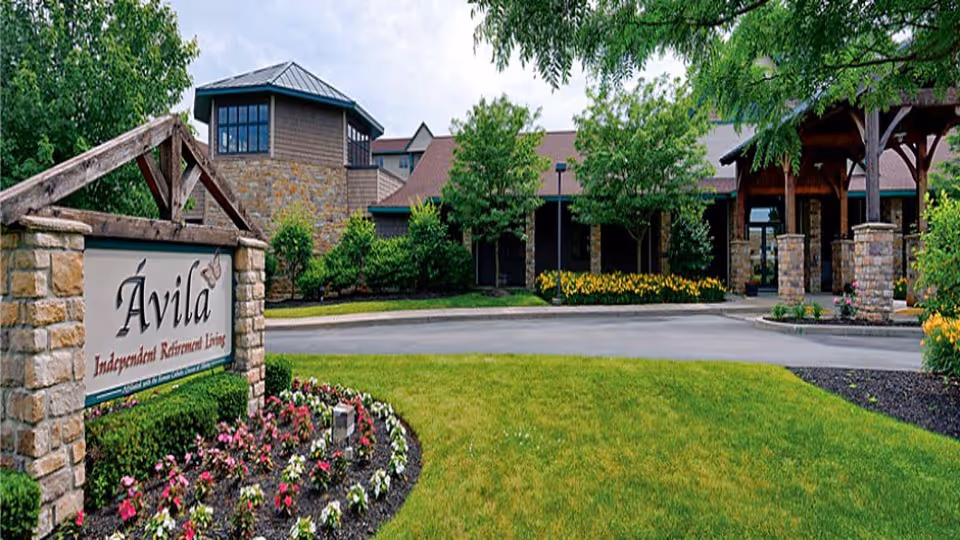 Entrance to Avila Independent Retirement Living community featuring a stone sign with the community name, surrounded by landscaped flower beds and green grass. The background shows a building with stone and wood exterior, trees, and a covered driveway.