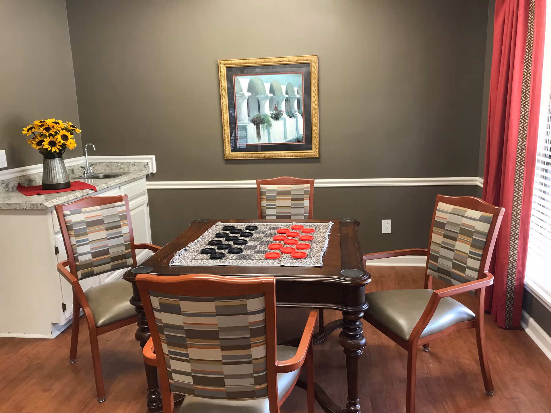 A cozy game room with a wooden table set for checkers, surrounded by four patterned chairs. A small countertop with a vase of sunflowers is visible on the left, and a framed painting hangs on the wall. Red curtains frame a window on the right, letting in natural light.