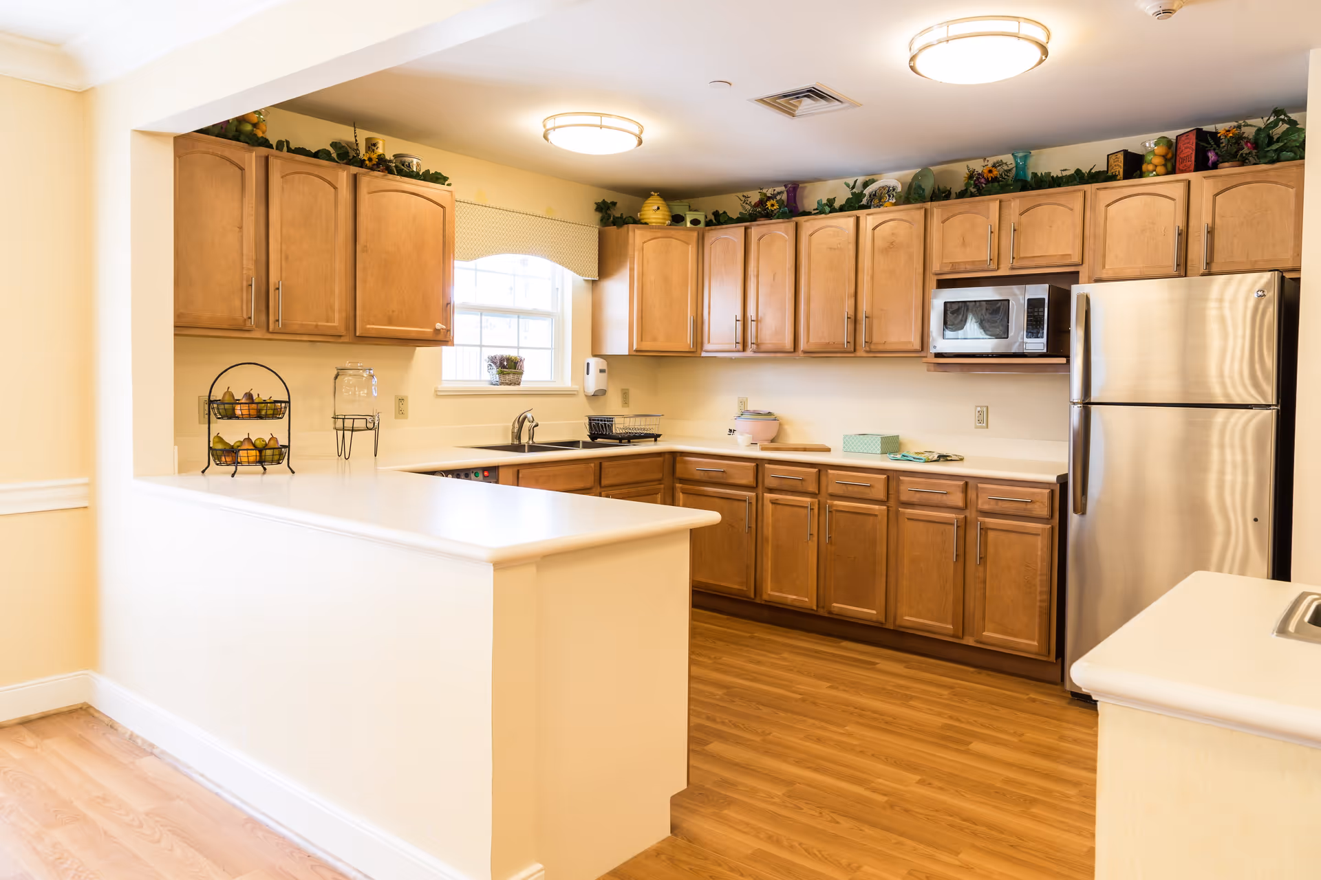 A bright kitchen with wooden cabinets, a stainless steel refrigerator, a microwave, a sink under a window, and a white countertop with a small fruit basket and a water dispenser.