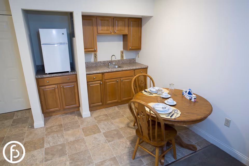 Small kitchen area with wooden cabinets, a countertop with a sink, and a white refrigerator. Next to the kitchen is a round wooden dining table set for two with plates, cups, and utensils. The floor is tiled and the walls are plain white.