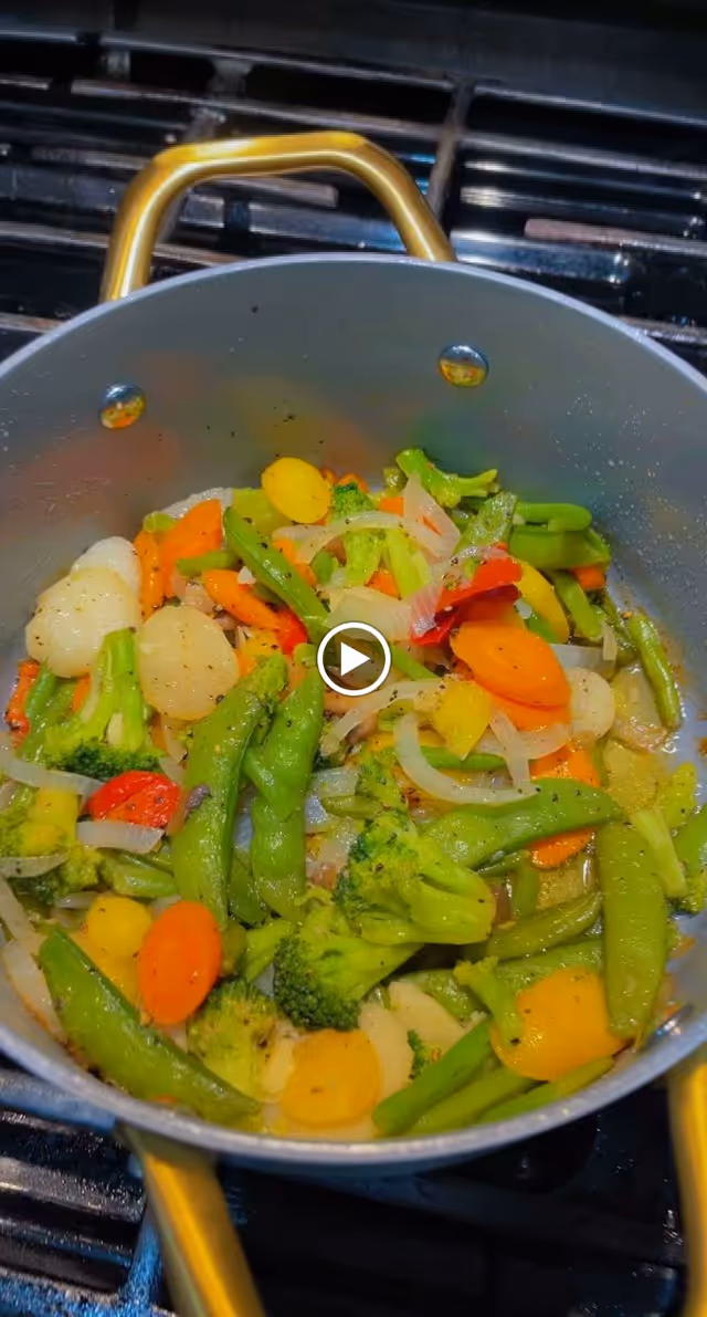 A pot on a stove filled with a colorful mix of cooked vegetables including broccoli, carrots, snap peas, onions, and bell peppers.