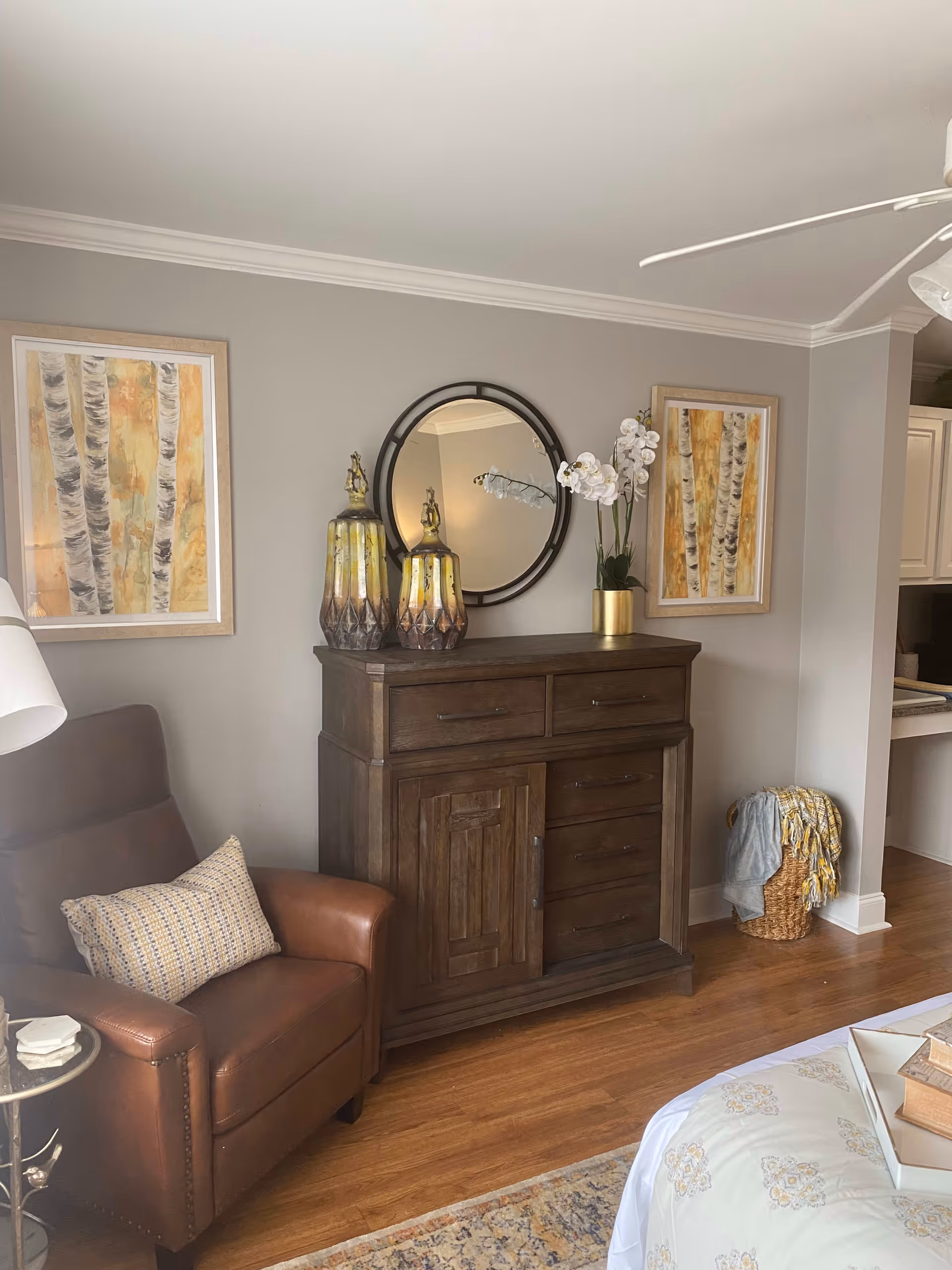 A cozy living area featuring a brown leather armchair with a patterned cushion, a dark wooden cabinet with drawers and doors, two decorative vases, a round wall mirror, and a potted white orchid. The walls are painted light gray and adorned with two framed paintings of birch trees. The floor is wooden, and a woven basket with a blanket is placed near the cabinet. Part of a bed with a floral bedspread and a small side table with books are also visible.