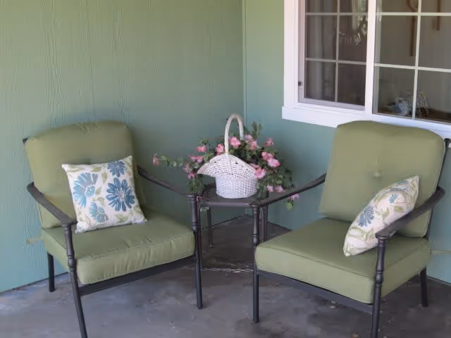Two green cushioned patio chairs with floral patterned pillows sit on a concrete floor next to a small glass-top table holding a white basket filled with pink flowers. The setting is against a green wall with a window.