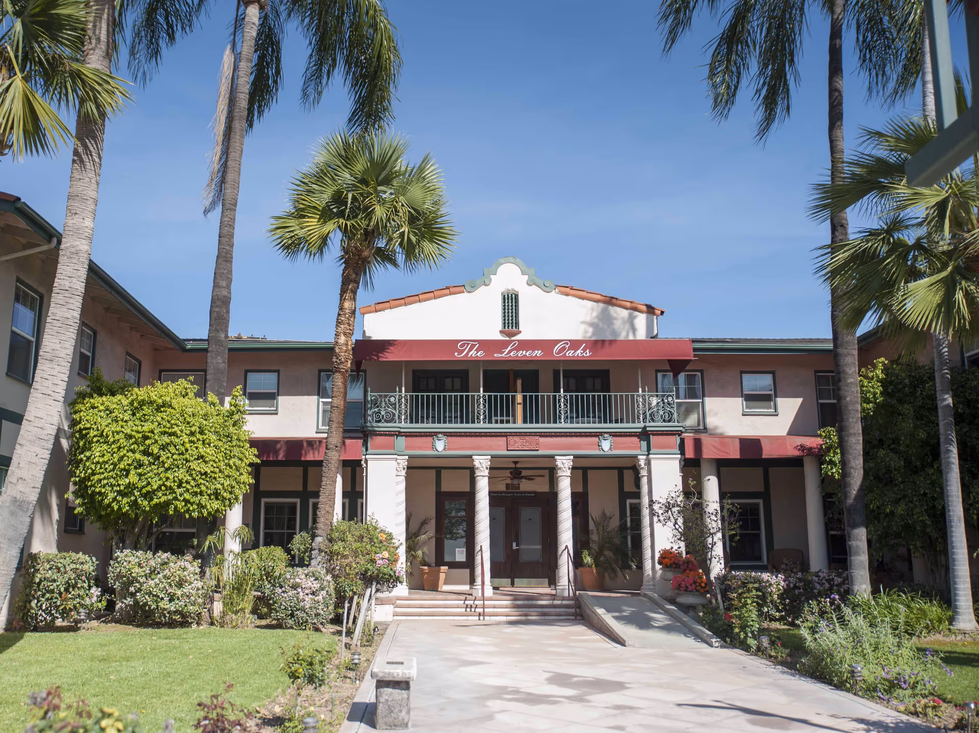 Front exterior view of a two-story building with a maroon awning that reads 'The Leven Oaks'. The building is surrounded by palm trees, bushes, and a well-maintained lawn under a clear blue sky.