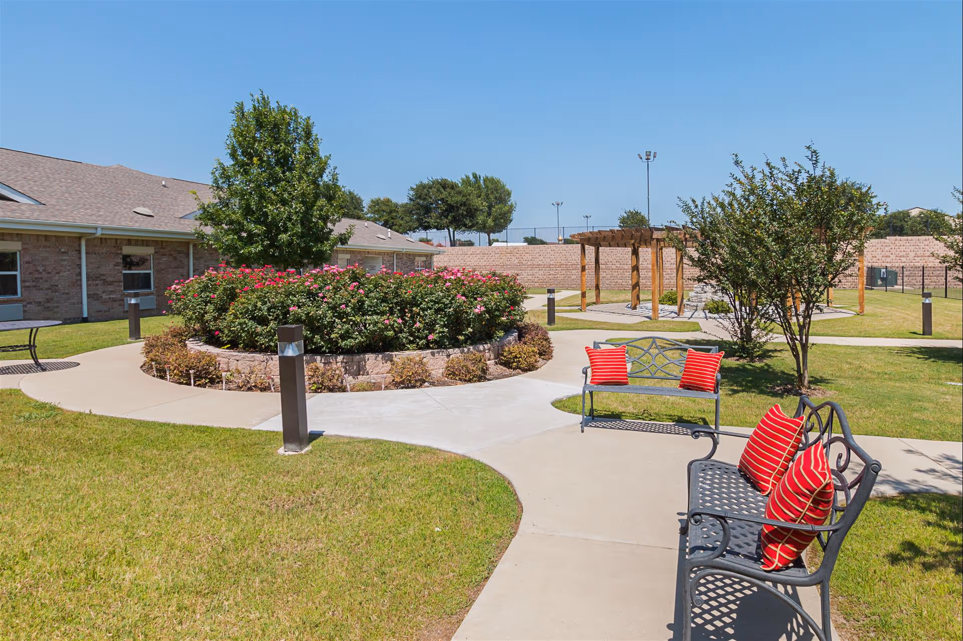 Outdoor garden area at Bethesda Gardens Fort Worth featuring a curved concrete walkway, green grass, a circular flower bed with blooming pink flowers, two black metal benches with red striped cushions, trees, and a wooden pergola in the background under a clear blue sky.