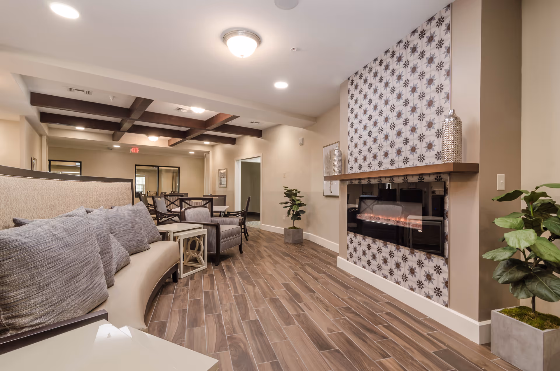 A cozy living room area in a senior living facility featuring a curved beige sofa with multiple gray cushions, a modern electric fireplace with decorative patterned tiles, wooden flooring, a small side table, several chairs, and potted plants. The ceiling has recessed lighting and wooden beams.