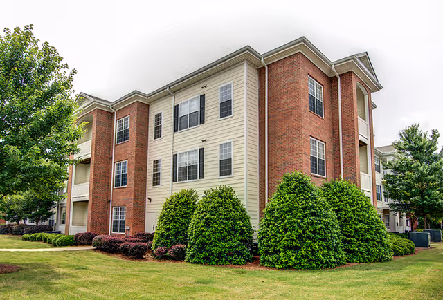 Exterior view of a three-story brick-and-siding apartment building with manicured lawn, shrubs, and trees.