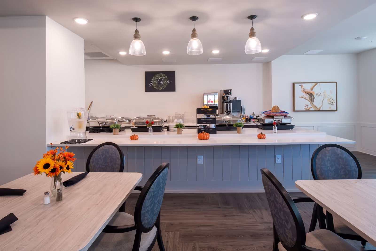 A dining area with light wood tables and cushioned chairs. The tables have black napkins and a vase with sunflowers. In the background, there is a buffet counter with chafing dishes, small decorative pumpkins, potted plants, and a beverage dispenser. Three pendant lights hang from the ceiling above the buffet. The walls are white with a framed bird artwork and a sign that says 'gather'.