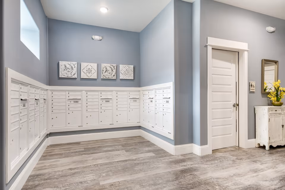 A clean and modern mailroom with multiple white mailboxes mounted on light blue walls. The room has wood-look flooring, a white door labeled as a women's restroom, a small white cabinet with a vase of yellow flowers, and decorative wall art above the mailboxes.