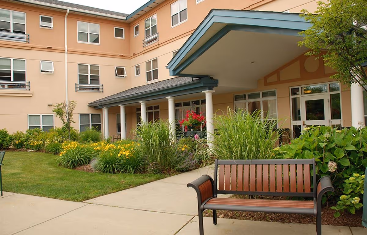 Front entrance of a residential building with a covered portico, landscaped flowerbeds and a bench on the sidewalk.