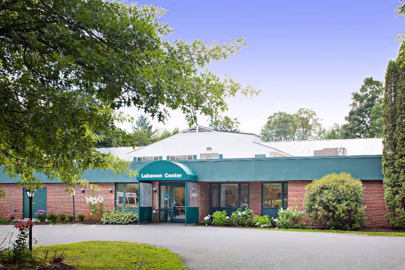 Exterior front view of Lebanon Center, a single-story brick building with a green awning over the entrance. The building is surrounded by trees and shrubs, with a paved driveway leading to the entrance.