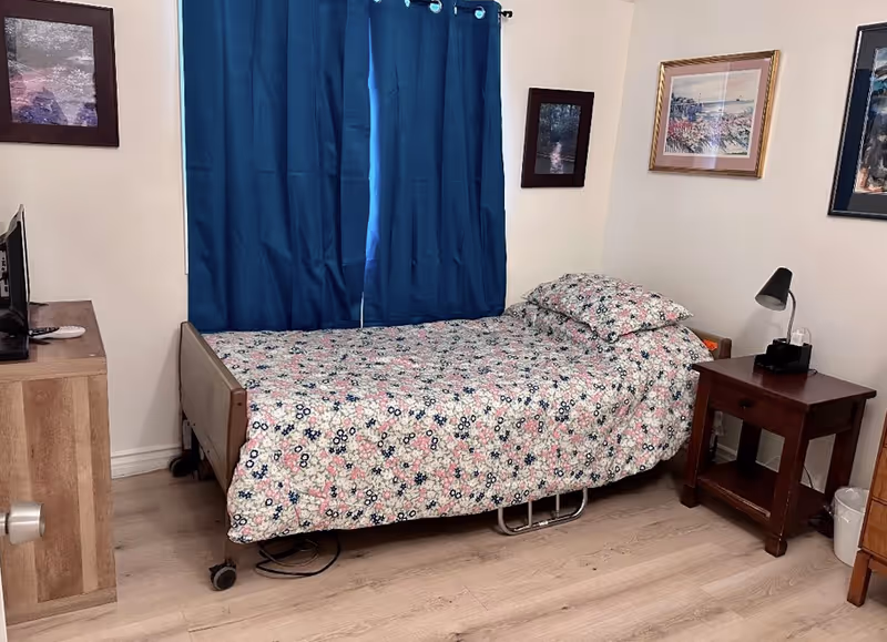 A small bedroom with a single bed covered in a floral patterned bedspread. The bed is positioned against a wall with blue curtains covering a window. There is a wooden nightstand with a lamp and some items on it next to the bed. The room has light-colored wooden flooring and framed pictures hanging on the walls. A wooden cabinet with a TV on top is visible on the left side of the image.