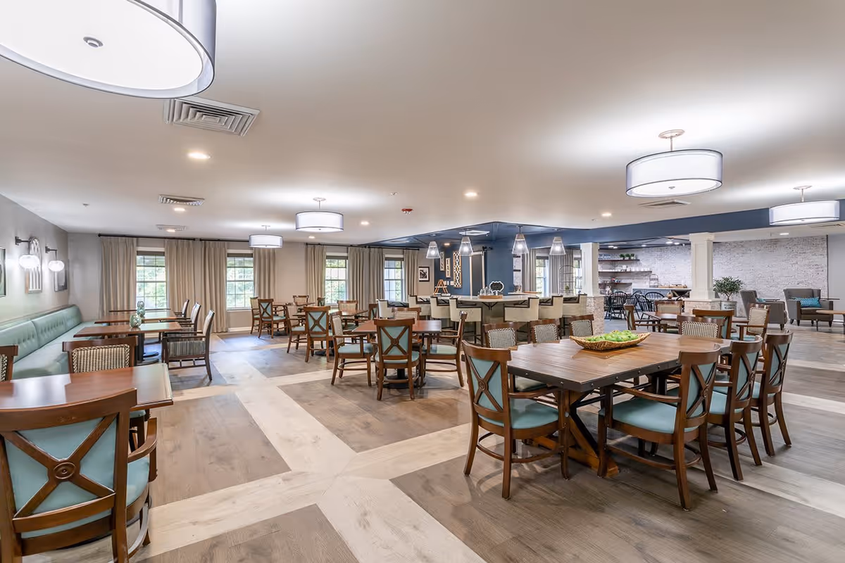 A spacious dining room with multiple wooden tables and chairs arranged neatly. The room features large windows with beige curtains, modern ceiling lights, and a combination of light and dark wood flooring. There is a bar area with high chairs and pendant lights, and a cozy seating area with armchairs in the background.
