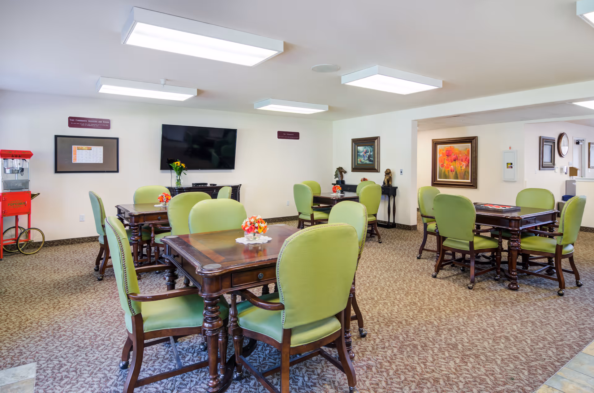 A bright and spacious common area with several wooden tables surrounded by green upholstered chairs. Each table has a small floral centerpiece. The room has carpeted flooring, white walls with framed artwork, a flat-screen TV mounted on the wall, and a red popcorn machine in the corner.