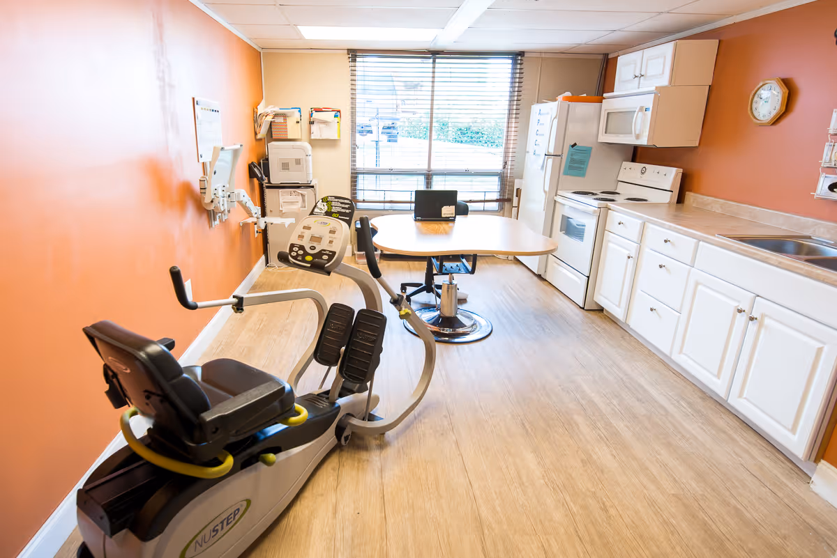 A room with an exercise bike in the foreground, a table with chairs near a large window with blinds, and a kitchen area with white cabinets, a stove, microwave, refrigerator, and a sink. The walls are painted orange and beige, and there is a clock on the wall.