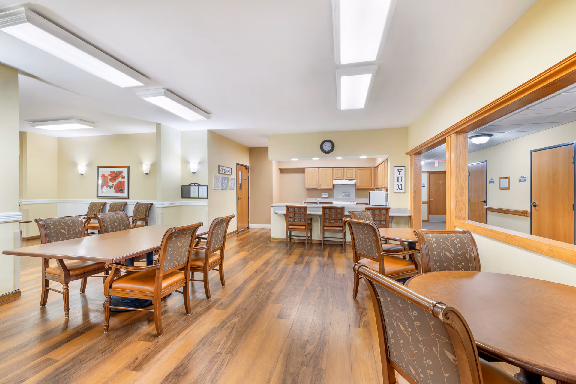 A bright dining area in a senior living facility with wooden tables and cushioned chairs. The room features wood flooring, a small kitchen area with cabinets, a stove, and a refrigerator. Wall decorations include a framed floral picture, a clock, and signs that say 'Let's eat' and 'YUM'. There is a large mirror on one wall reflecting the hallway with wooden doors.