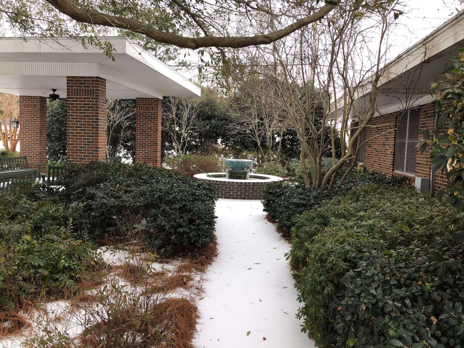 Outdoor garden area at Naomi Heights Nursing and Rehabilitation Center, LLC featuring a white pathway leading to a circular brick water fountain surrounded by bushes and trees. There is a covered seating area with brick pillars and green benches on the left side.