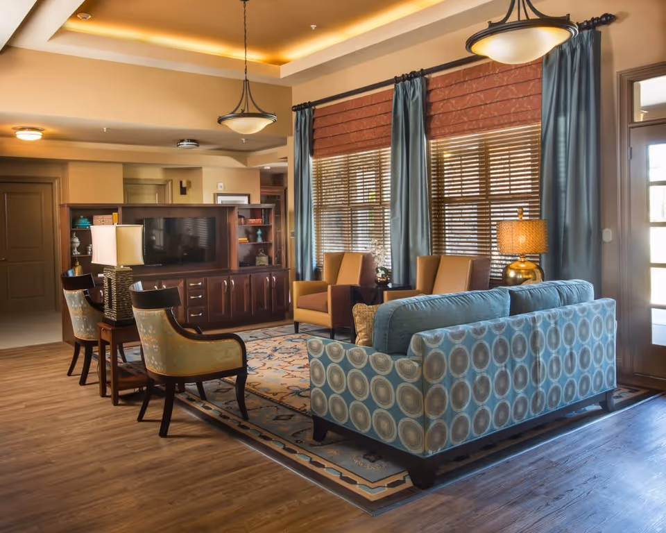 A cozy living room area in an assisted living facility featuring a patterned blue sofa, two brown armchairs, two upholstered chairs with floral patterns, a wooden TV cabinet with a flat-screen TV, a table lamp, large windows with wooden blinds and blue curtains, and warm lighting from ceiling fixtures.