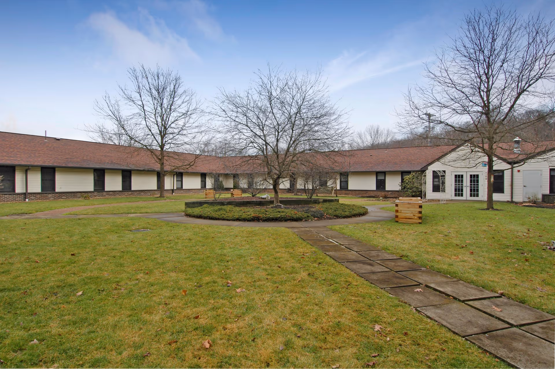 Outdoor courtyard area of a senior living facility with a circular garden bed in the center, surrounded by leafless trees and a paved walkway leading to the building. The building has a beige exterior with a brown roof and multiple windows.
