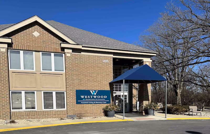Exterior view of a brick building with a blue canopy over the entrance. The building has multiple windows and a sign that reads 'Westwood Riverside Senior Life Communities Assisted Living & Memory Care'. There are some trees and benches visible on the right side under a clear blue sky.
