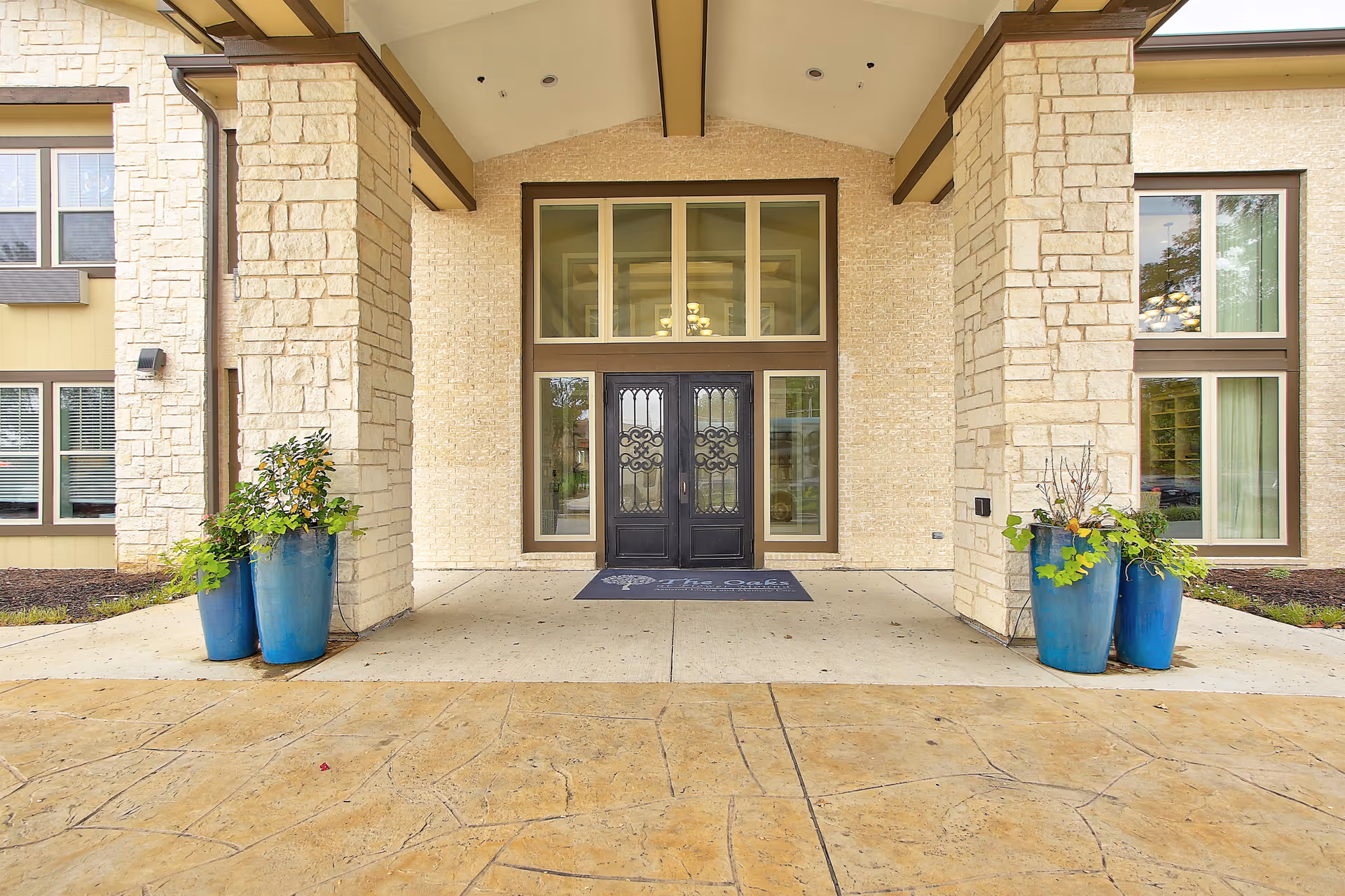 Entrance of The Oaks at Flower Mound facility featuring double black decorative doors framed by large windows, stone pillars on either side, and blue planters with green plants.