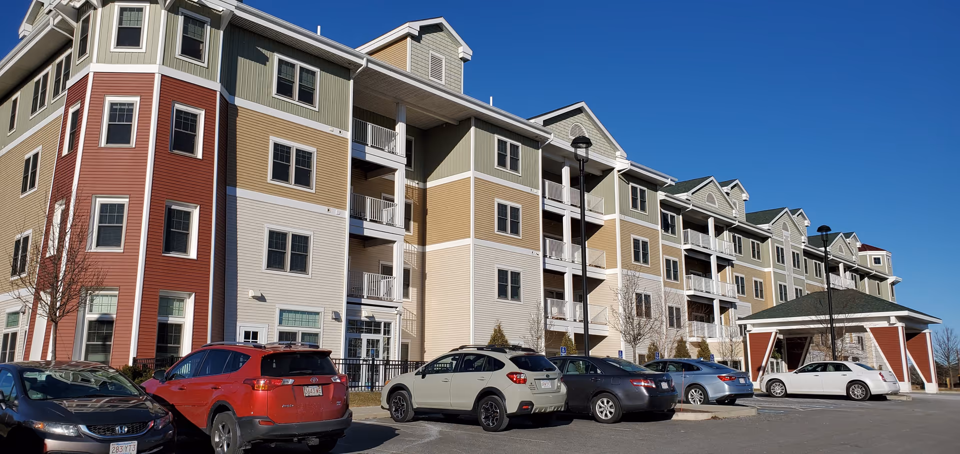 Exterior view of a multi-story senior living facility building with beige, green, and red siding. Several cars are parked in front of the building under a clear blue sky.