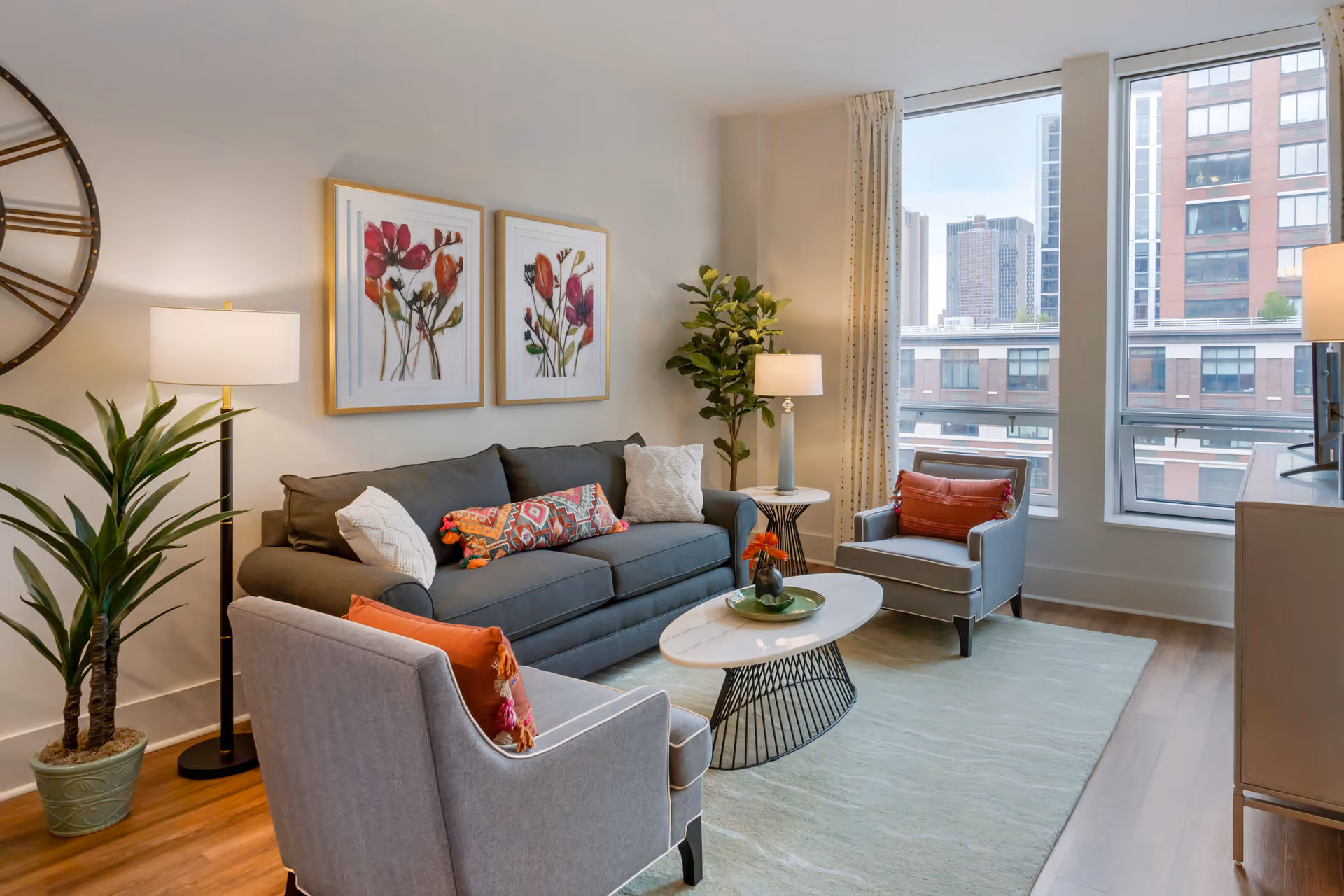 A modern living room with a gray sofa and two gray armchairs, each with colorful cushions. There is a round white coffee table with a decorative item on it, a large wall clock, two framed floral artworks, two floor lamps, and two potted plants. Large windows show a cityscape outside.