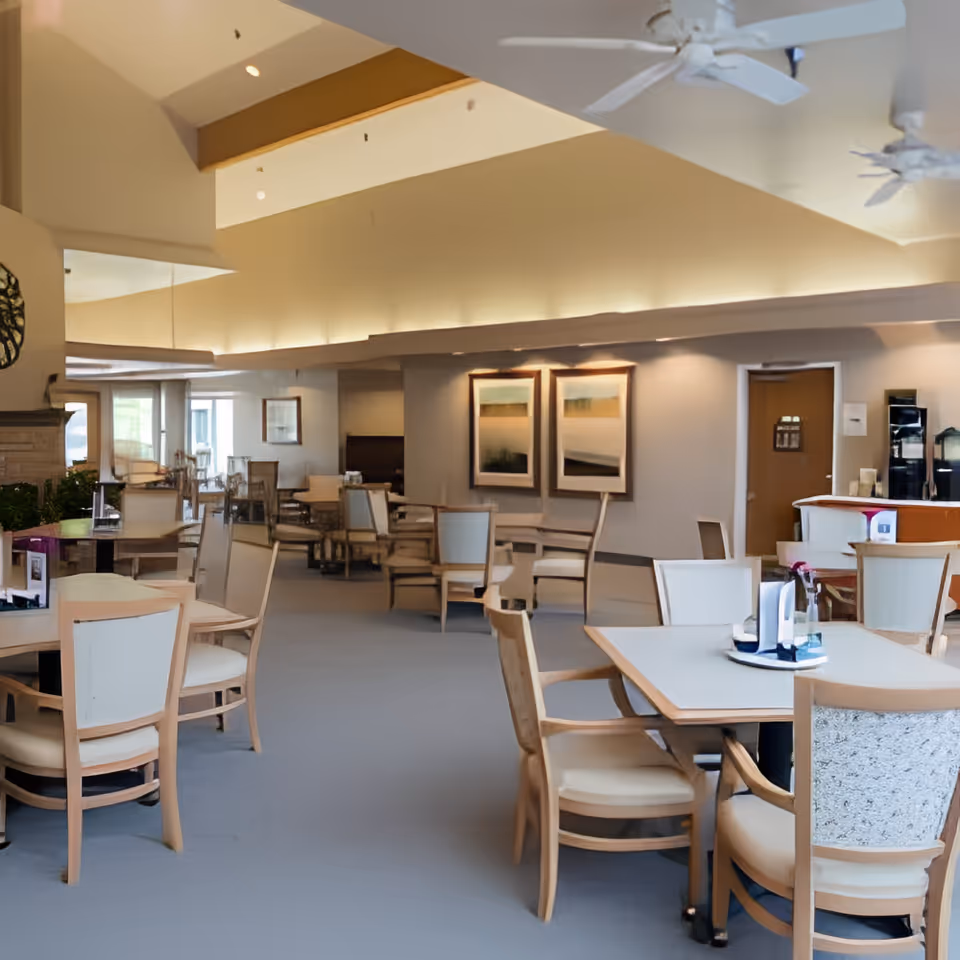 A spacious dining room with multiple tables and chairs arranged neatly. The room features a high ceiling with ceiling fans, soft lighting, and framed artwork on the walls. The decor is warm and inviting, suitable for communal dining.