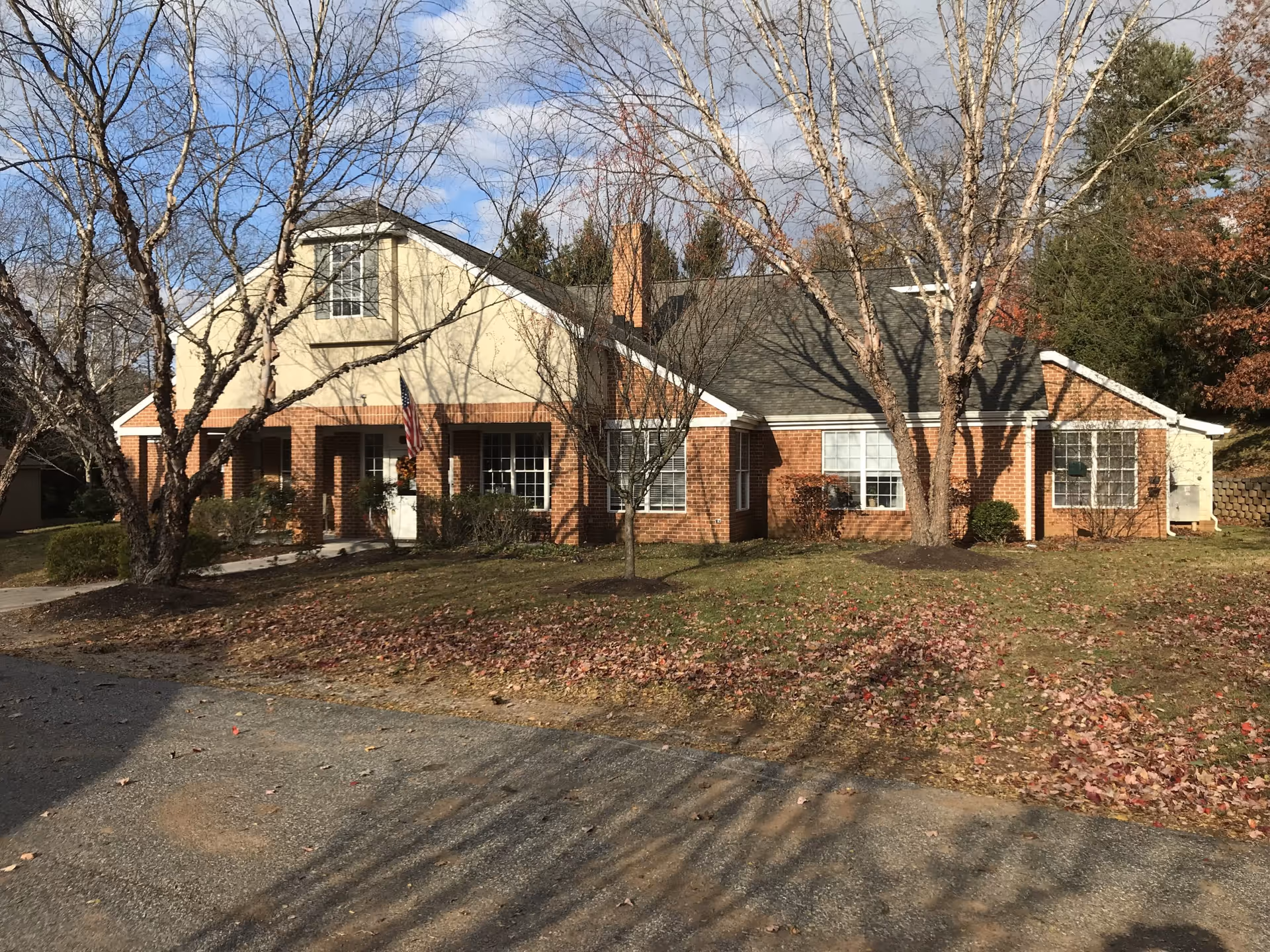 Exterior view of a single-story brick and siding building with a gray shingled roof, several windows, and a front entrance with an American flag. Leafless trees and some fallen leaves are visible on the lawn and driveway in front of the building under a partly cloudy sky.