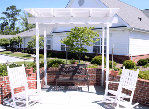 Outdoor seating area with two white rocking chairs and a black metal bench under a white pergola. The area is surrounded by low brick walls with potted plants, and a small tree is planted behind the bench. In the background, there is a white building with a gray roof and some greenery.
