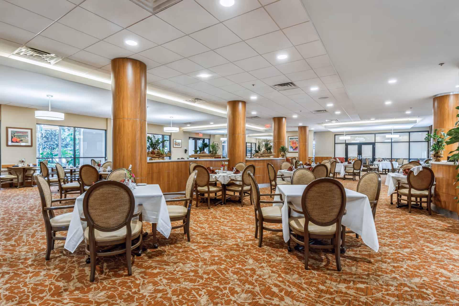 A spacious dining room with multiple tables covered in white tablecloths and surrounded by cushioned wooden chairs. The room features large wooden columns, patterned carpet, ceiling lights, and windows letting in natural light. There are plants and framed pictures on the walls, creating a warm and inviting atmosphere.