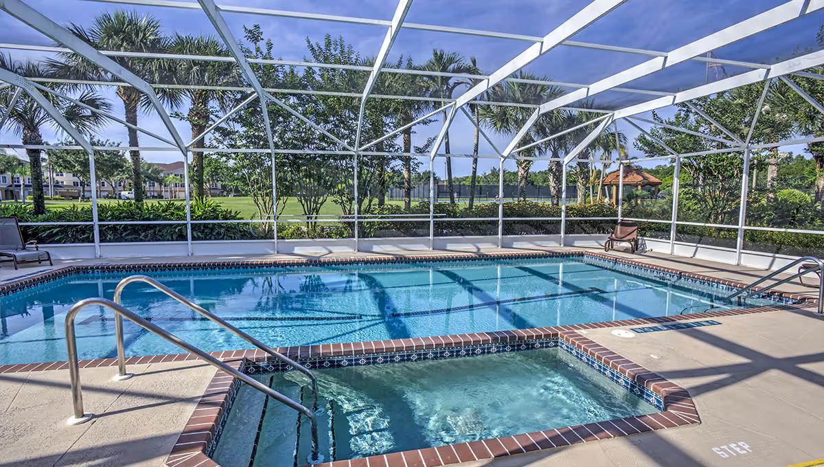 A screened-in outdoor swimming pool and hot tub area with metal handrails, surrounded by a concrete deck with lounge chairs. Beyond the enclosure, there are palm trees, bushes, and a grassy area under a clear blue sky.